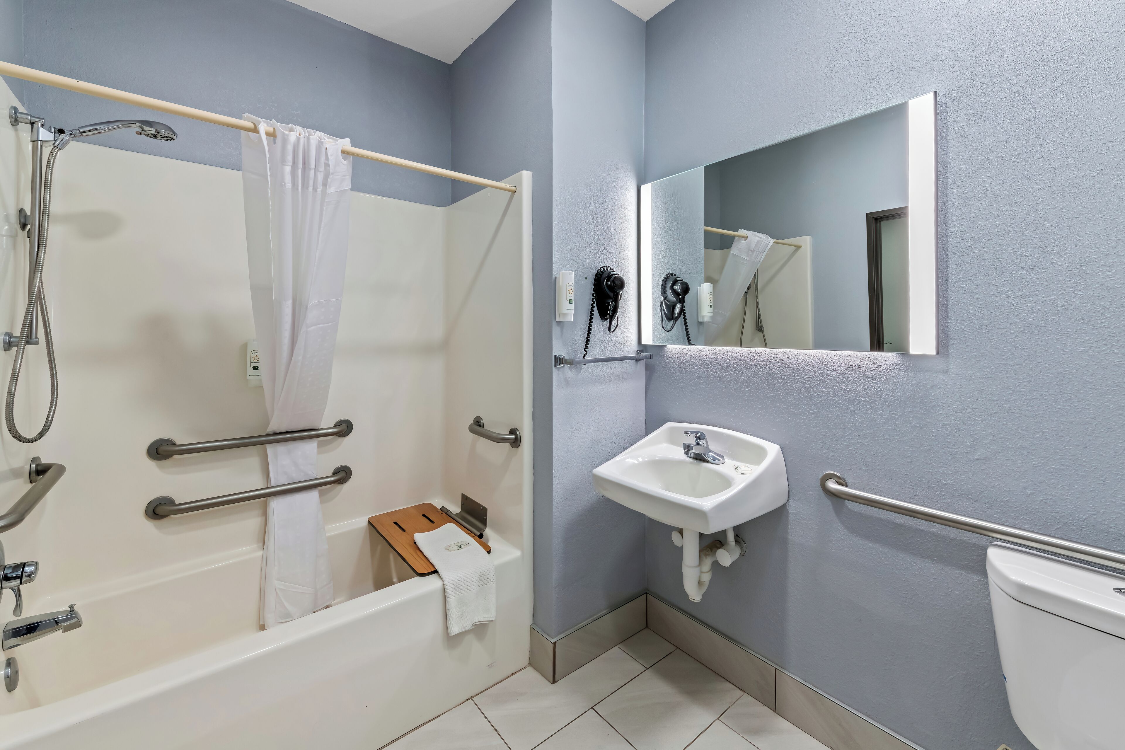 A modern bathroom with a bathtub, shower, and sink, equipped with handrails and a mirror.