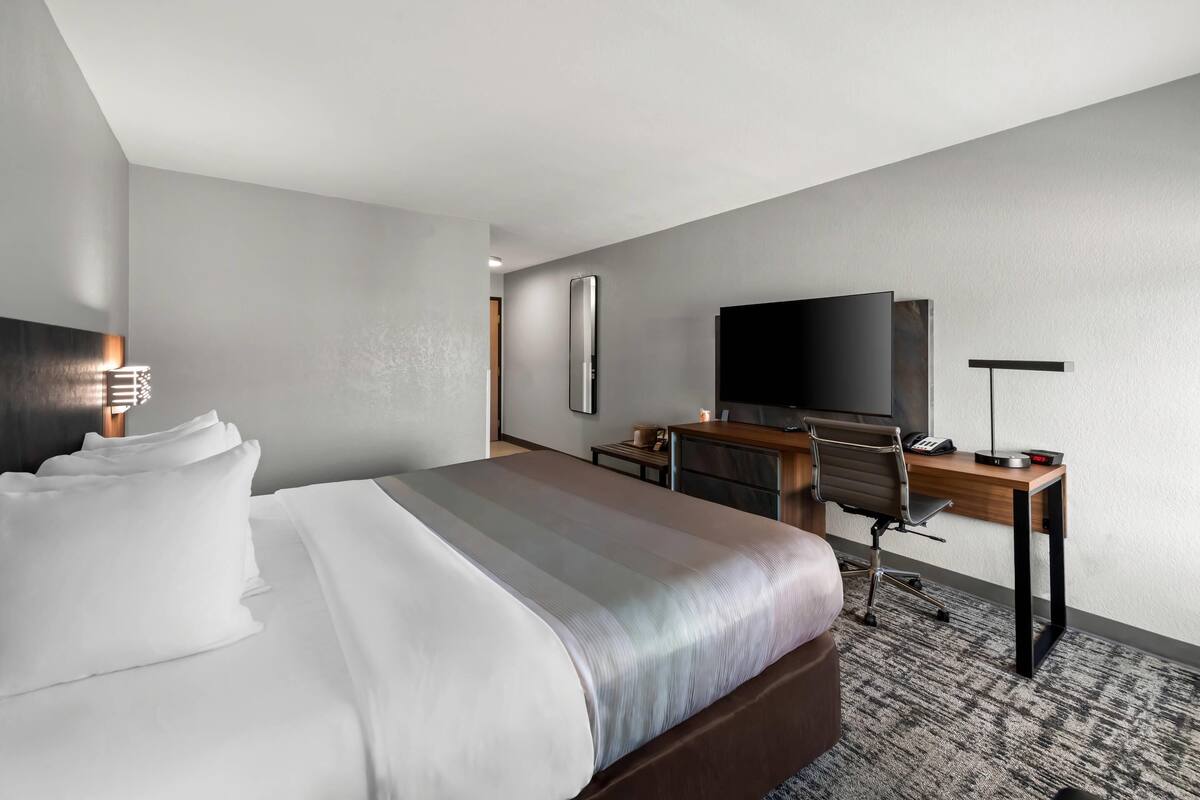 A hotel room with a large bed, a desk with a chair, a flat-screen TV, and a bedside table with a lamp.