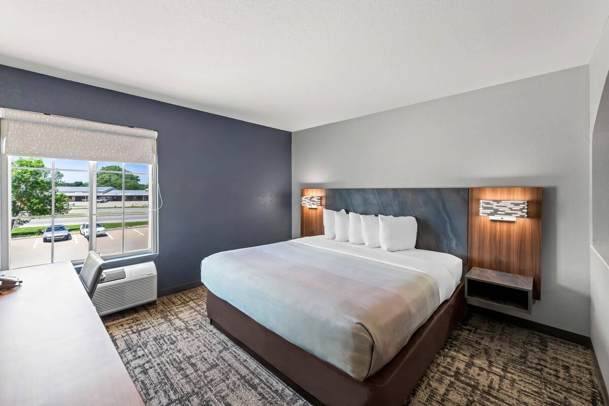 Ein Hotelzimmer mit einem großen Bett, einem Schreibtisch und einem Fenster mit Blick auf einen Parkplatz und Bäume.