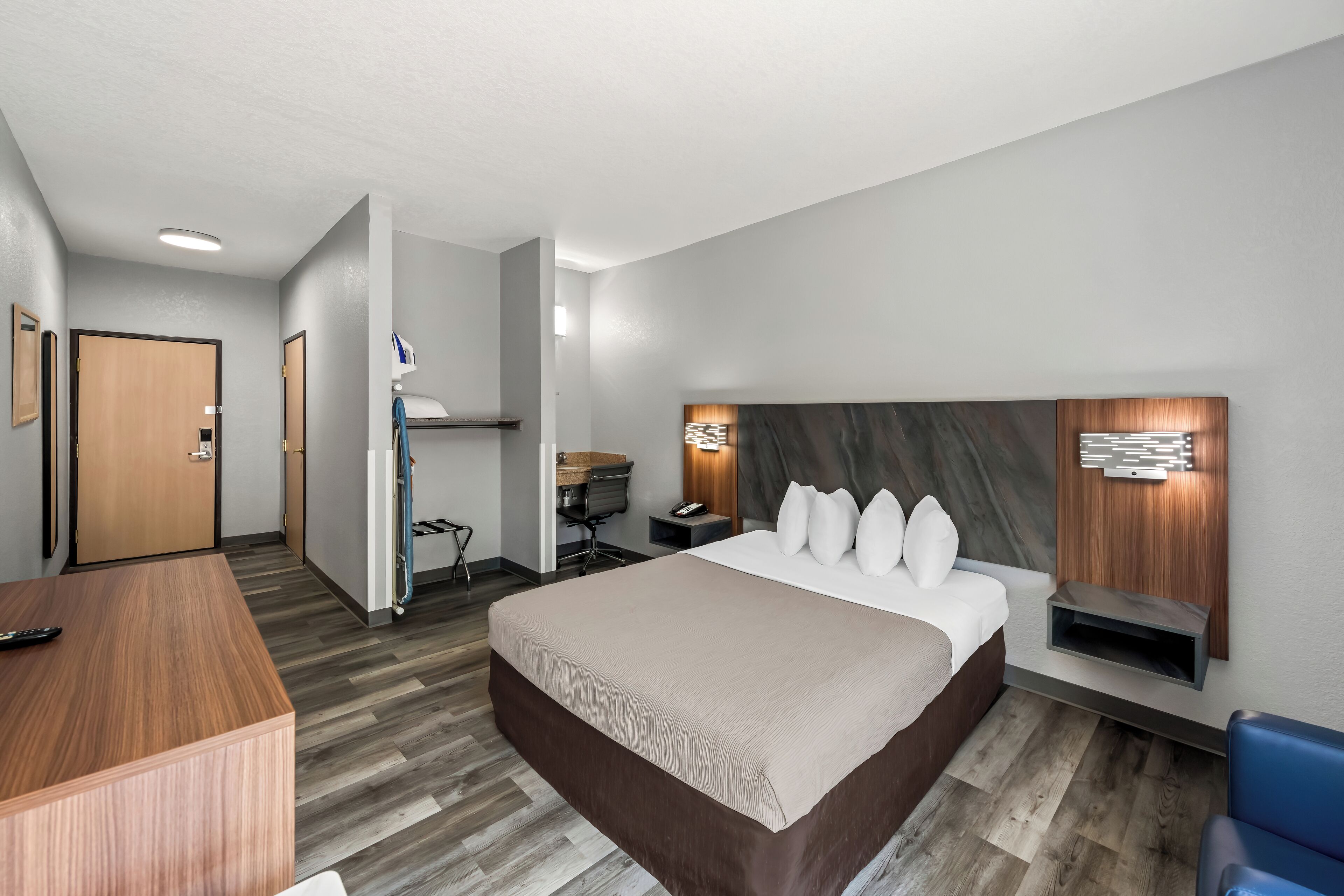 A modern hotel room with a large bed, a desk, a chair, and a small seating area with a wall-mounted shelf.
