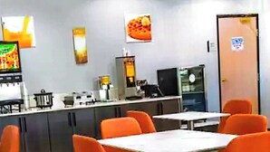 A dining area with orange chairs and white tables, a kitchen in the background, and a door with a sign.