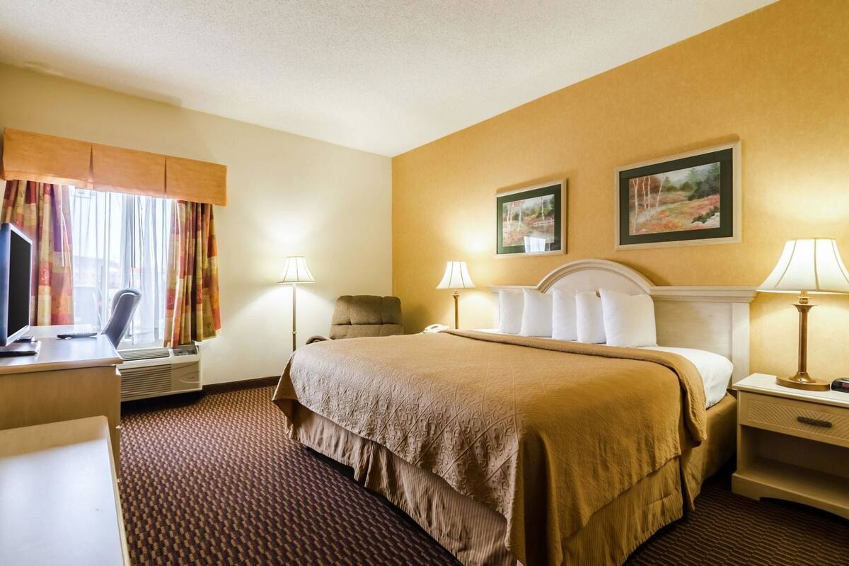 Ett hotellrum med en säng, ett skrivbord, en stol, två sänglampor, en platt-TV och två inramade tavlor på väggen.