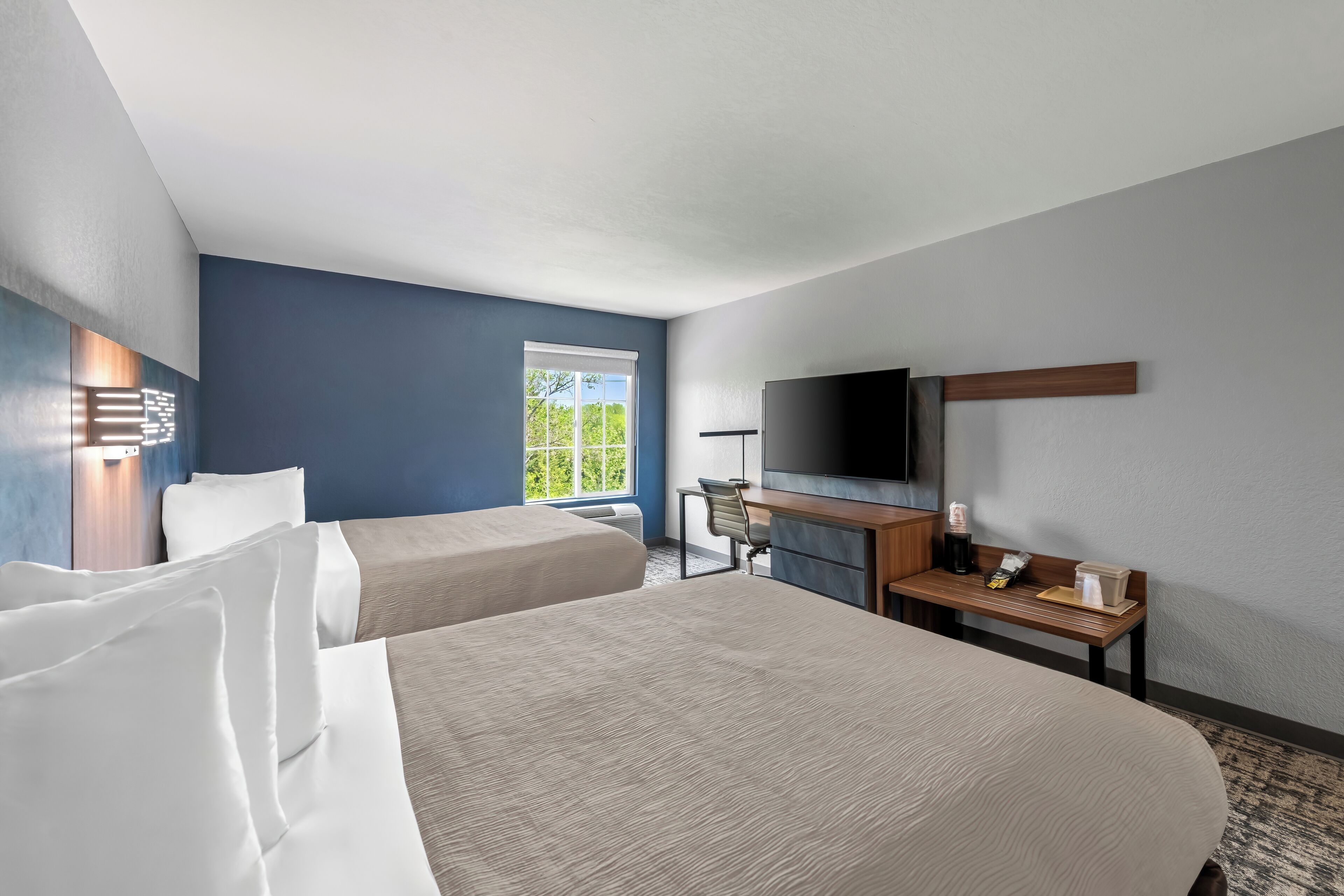 A hotel room with two beds, a desk with a chair, a flat-screen TV, and a window with a view of greenery.