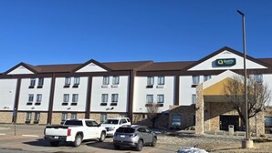 A multi-story hotel with a central entrance, parking lot, and a clear blue sky.