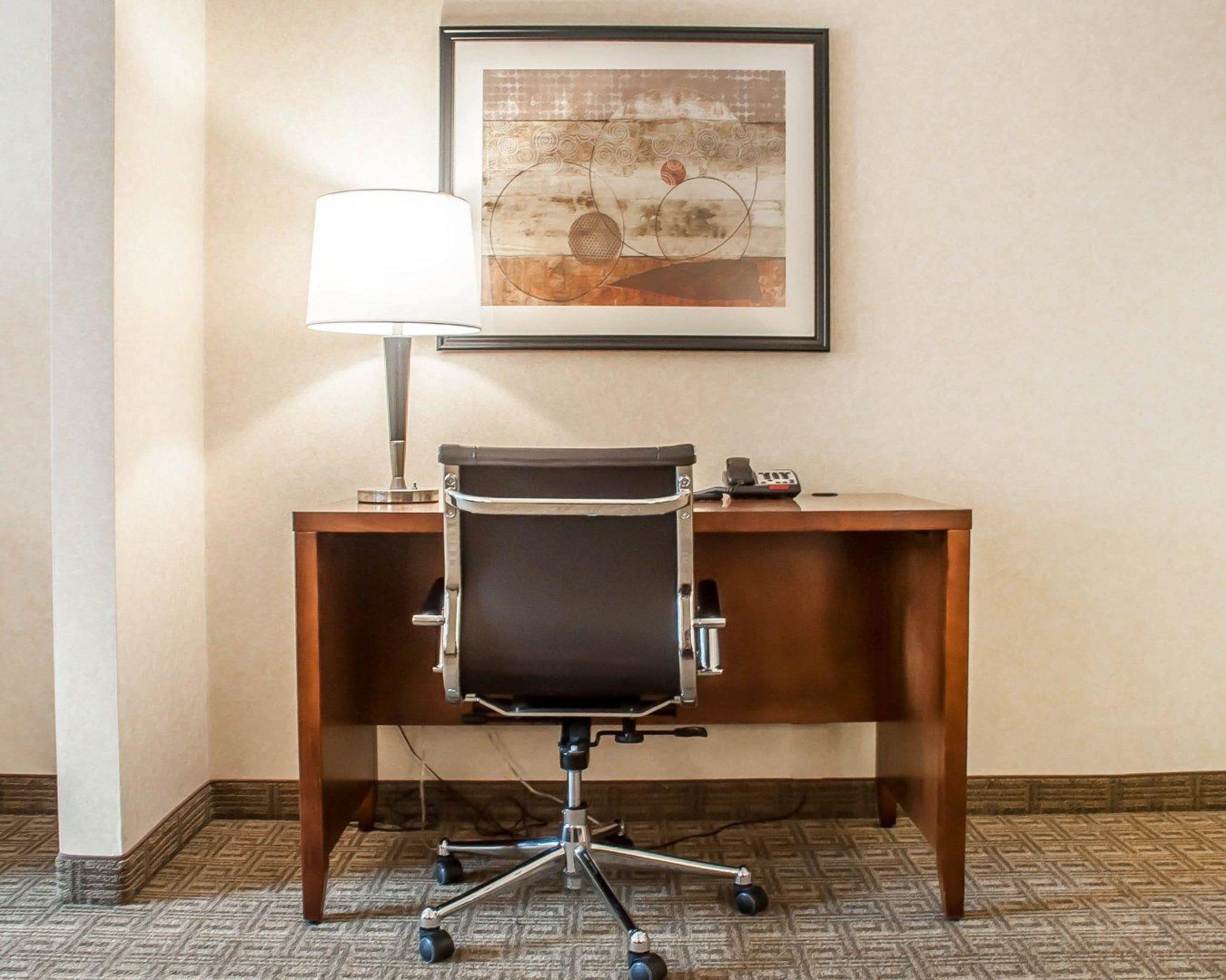 A hotel room with a wooden desk, a black office chair, a lamp, and a framed abstract painting on the wall.