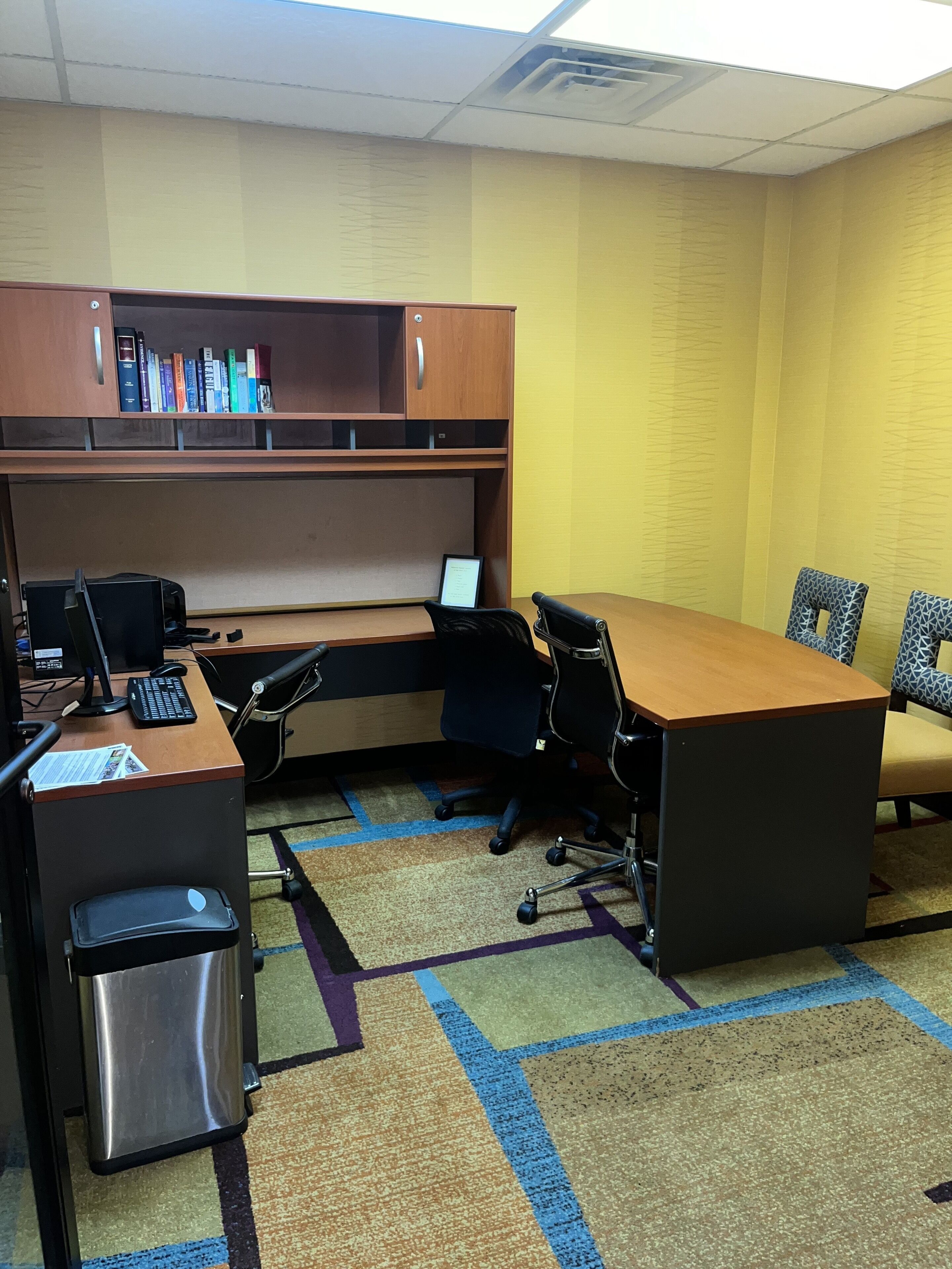 A conference room with a long table, chairs, a desk with a computer, a bookshelf, and a trash bin.