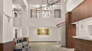 Um lobby moderno de hotel com teto alto, um lustre central, um relógio na parede e uma área de estar com poltronas.
