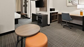 Quarto de hotel com uma cama, uma escrivaninha com computador, televisão, micro-ondas e uma pequena mesa com um banquinho laranja.