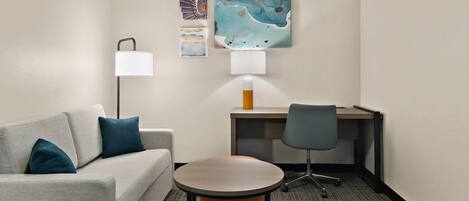 A hotel room with a grey sofa, a round table, a desk, and two lamps.