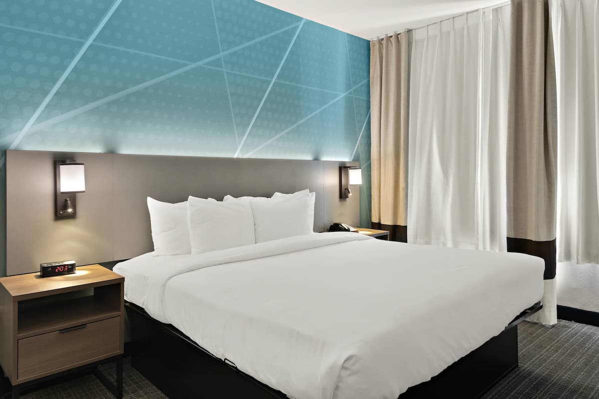 Una habitación de hotel con una cama grande, una mesita de noche con lámpara y un reloj en la pared.