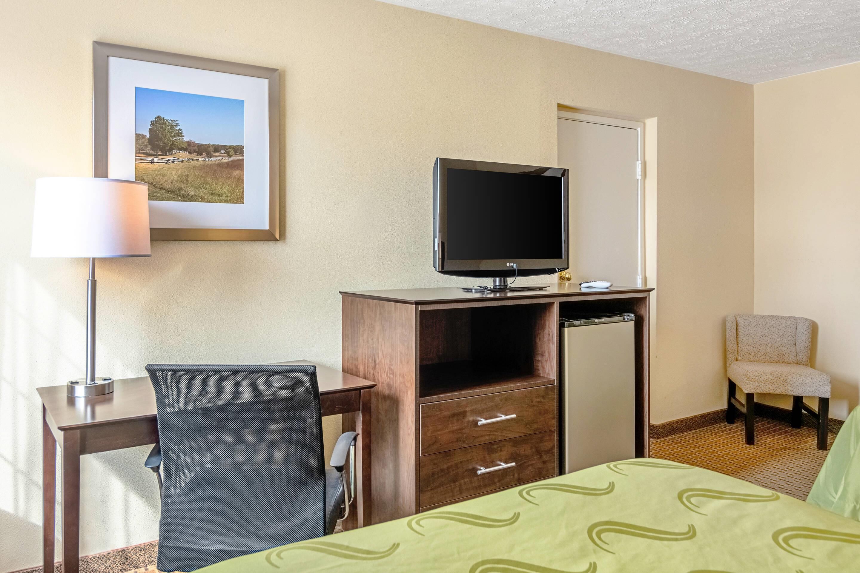 A hotel room with a bed, a desk with a chair, a television, a refrigerator, and a chair.