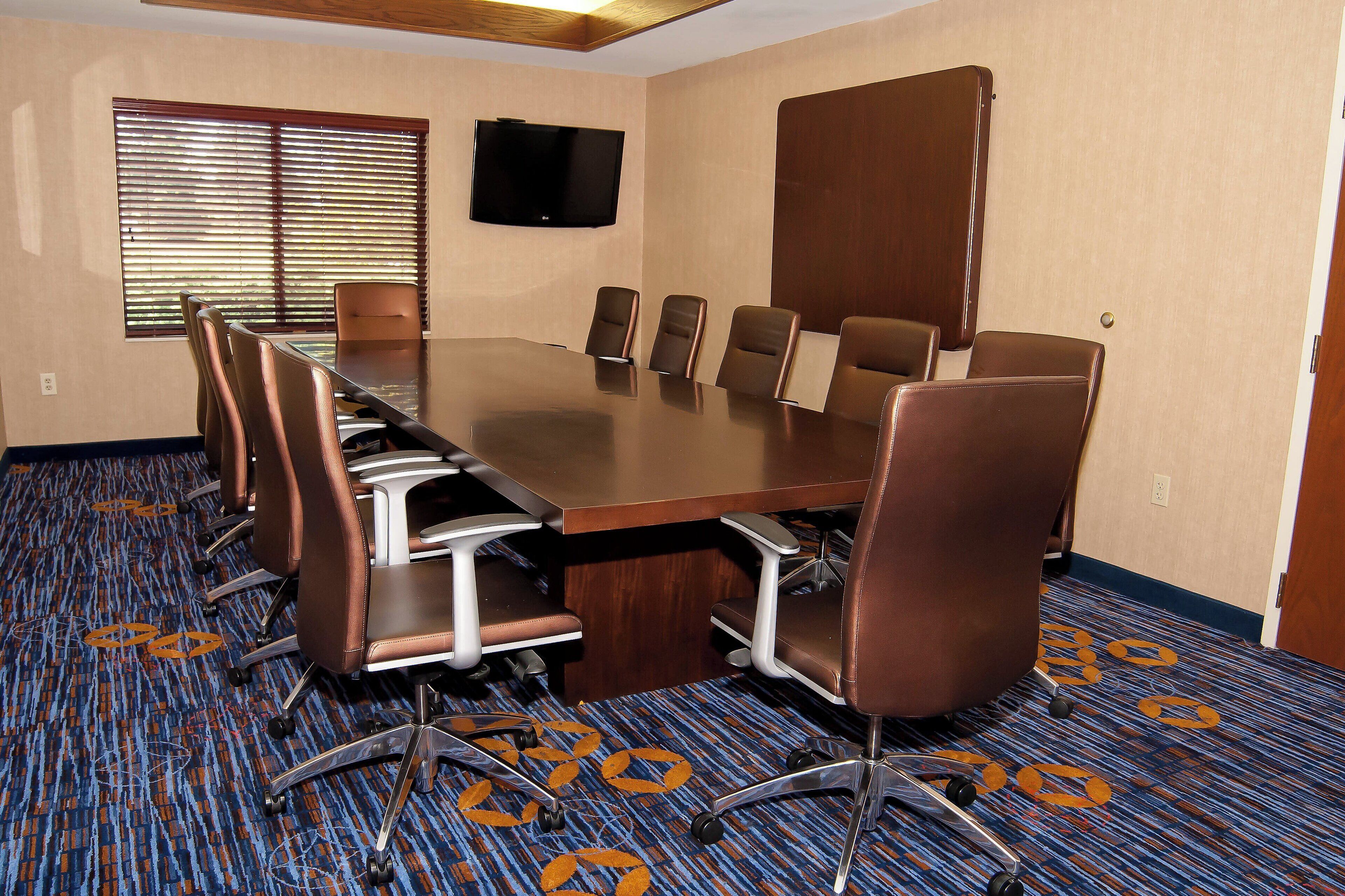 A conference room with a large table, six chairs, a television on the wall, and patterned carpet.