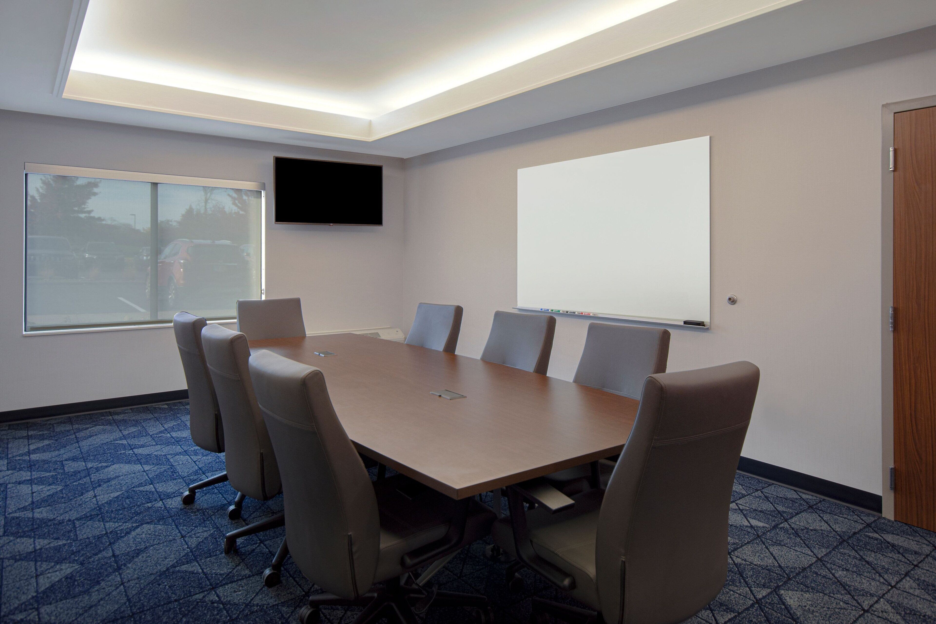 Un salón de conferencias con una gran mesa, seis sillas, una pantalla de proyector, un televisor y una puerta.
