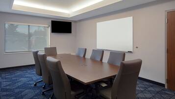 Un salón de conferencias con una gran mesa, seis sillas, una pantalla de proyector, un televisor y una puerta.