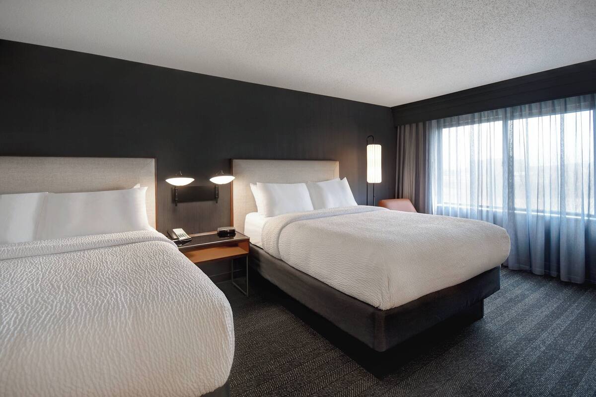 A hotel room with two beds, a nightstand with a lamp, a telephone, and a window with curtains.