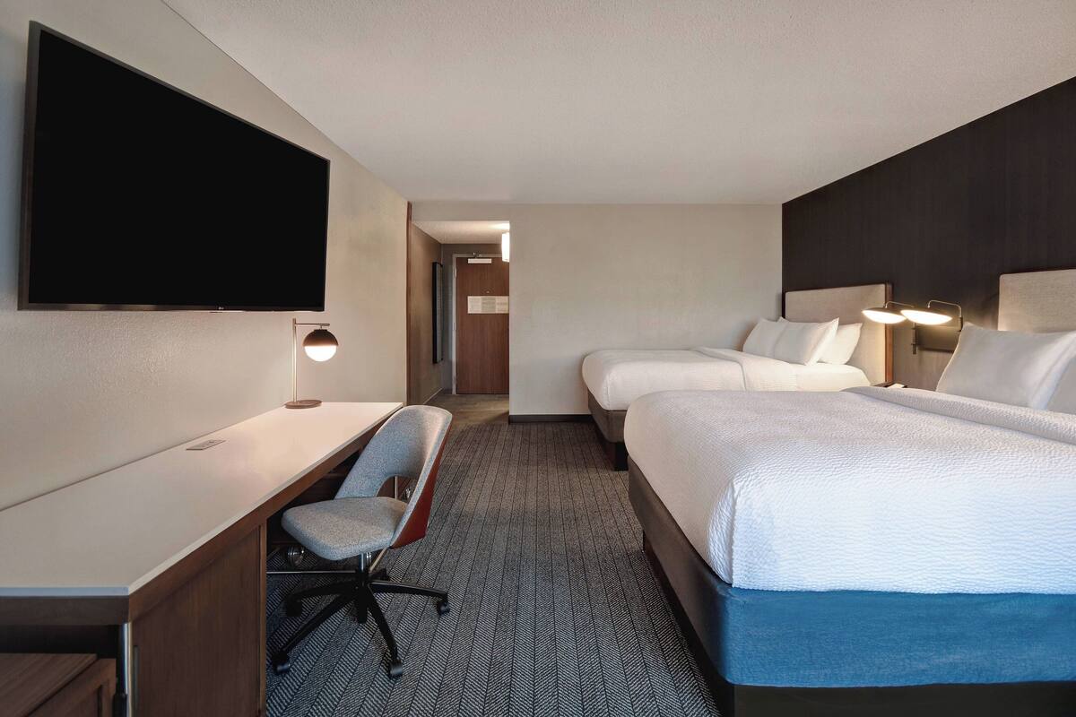 A hotel room with a flat-screen TV, a desk with a chair, two beds, and bedside lamps.