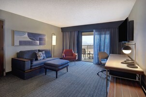 Habitación de hotel con sofá, reposapiés, escritorio, silla y una ventana grande con cortinas.