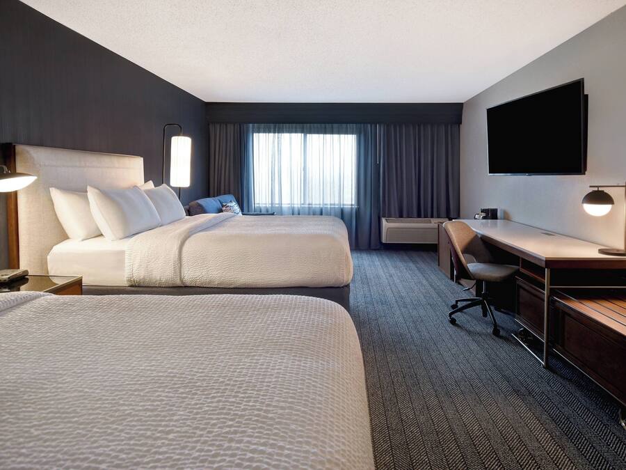 A hotel room with two beds, a desk, a chair, a flat-screen TV, and a window with curtains.