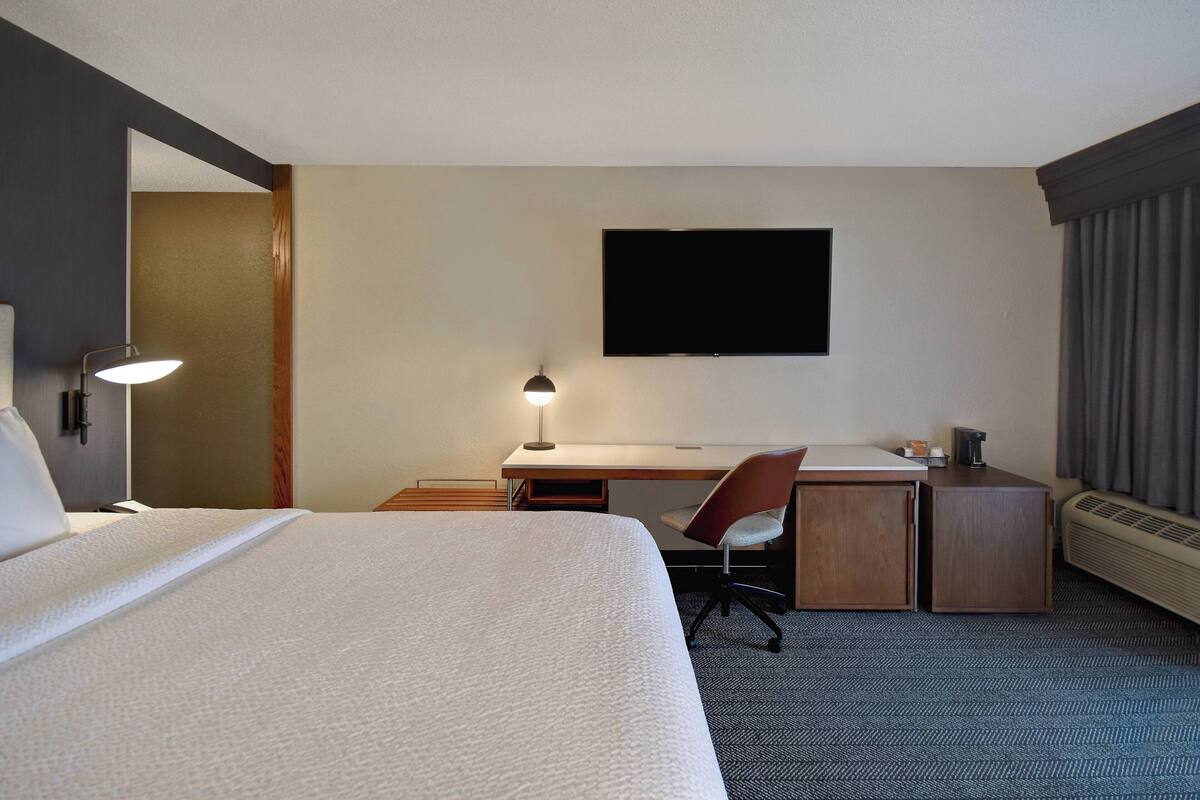 A hotel room with a large bed, a desk, a chair, a flat-screen TV, a lamp, and a heater.