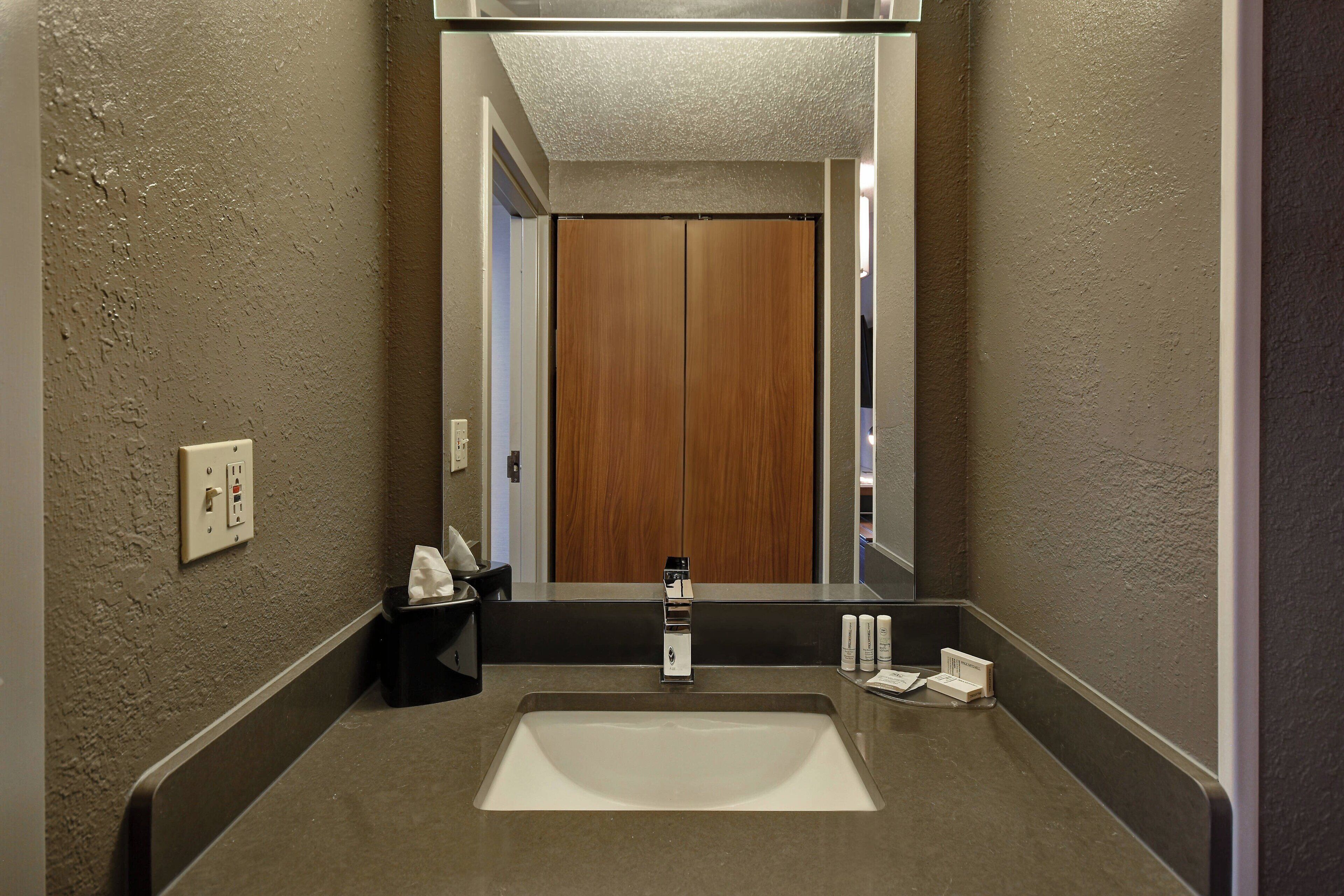 A bathroom with a large sink, a mirror, a wooden door, and a towel dispenser.