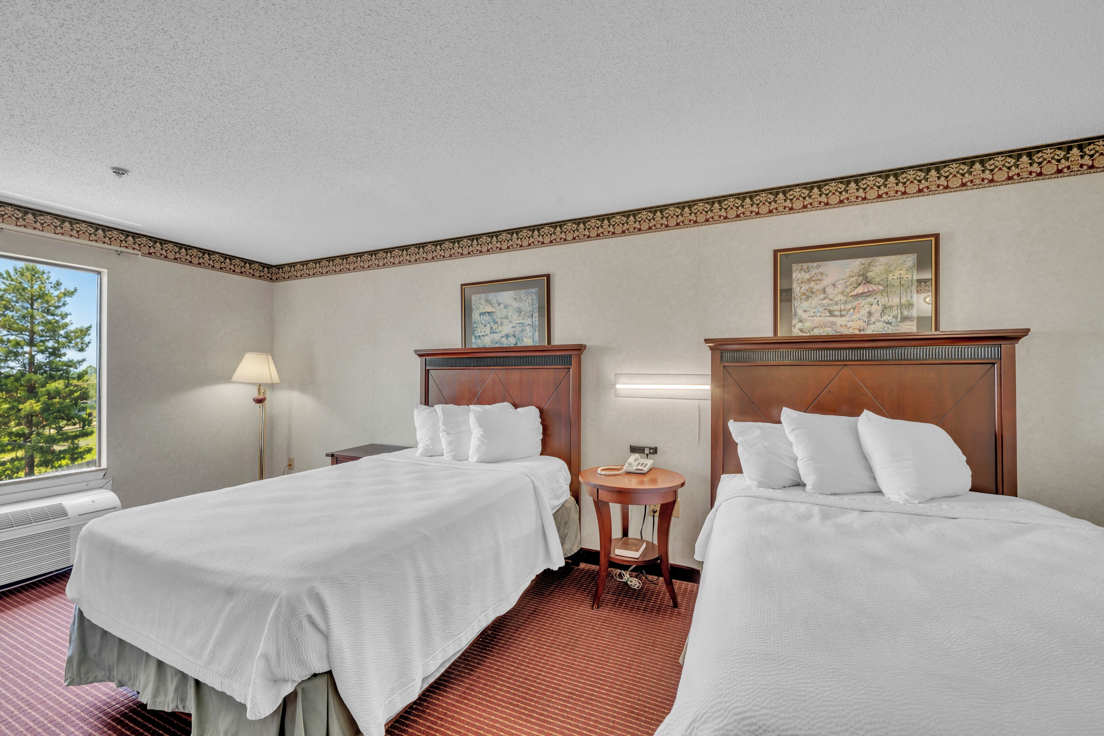 A hotel room with two beds, a bedside table with a lamp, a small round table with a phone, and a window with a view of trees.
