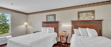 A hotel room with two beds, a bedside table with a lamp, a small round table with a phone, and a window with a view of trees.