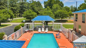A rectangular swimming pool with a blue roofed pavilion, surrounded by a white fence, and a view of a road and trees in the background. - Gray Inn and Suites (Gray)