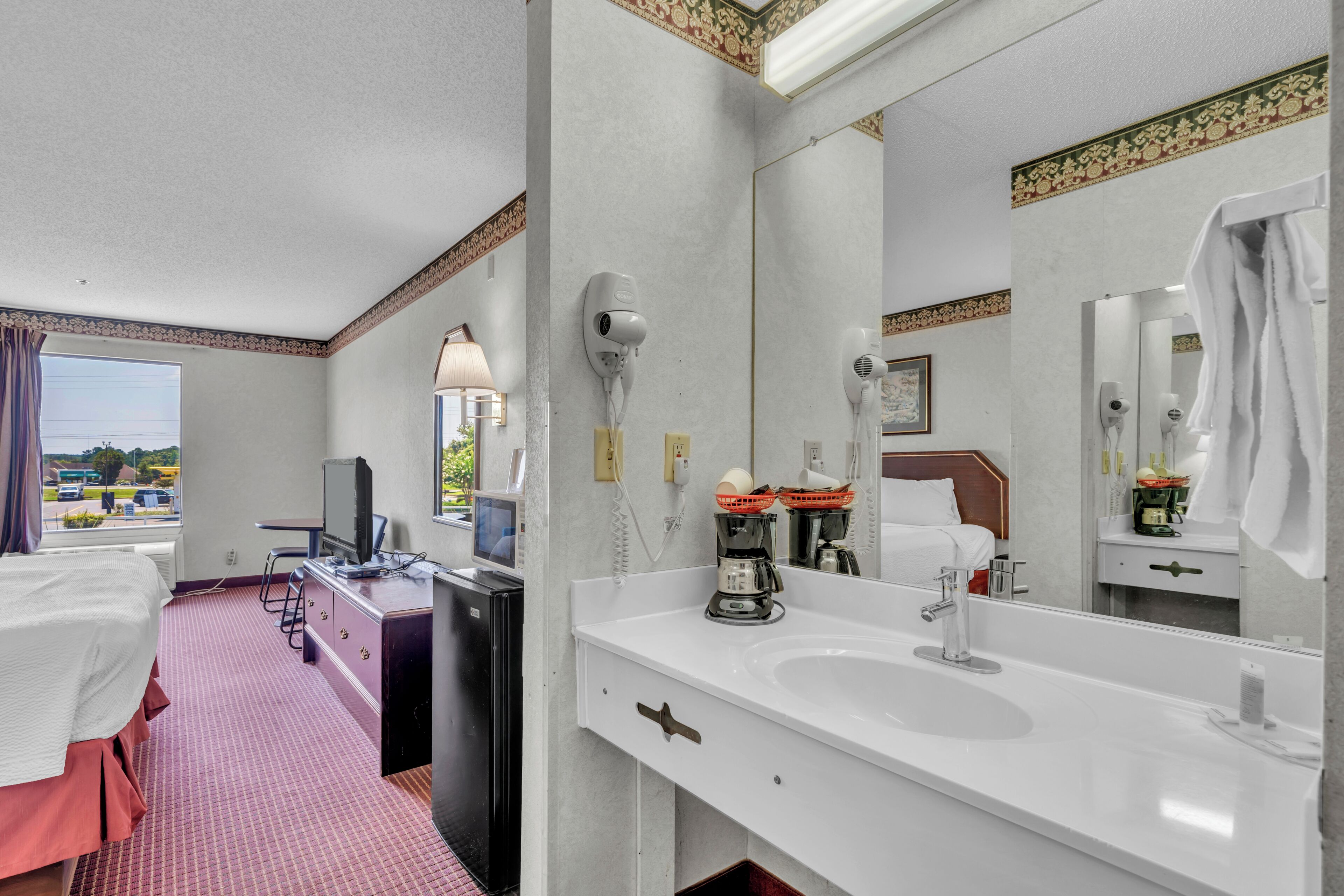 A hotel room with a bed, a desk with a computer, a large mirror, a sink, and a window with a view of the outside.