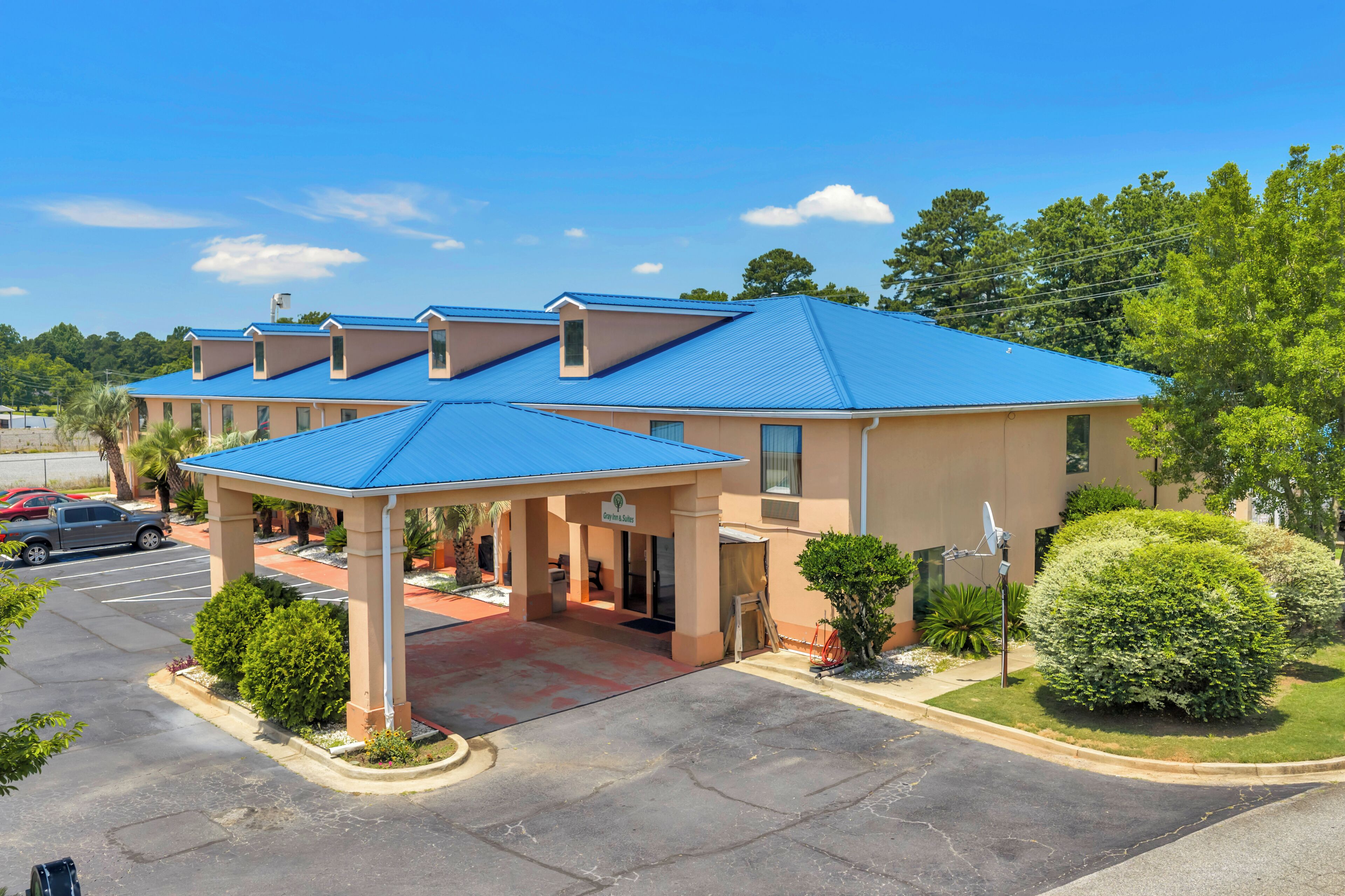A hotel with a blue roof, covered parking, and a landscaped area with bushes and trees.