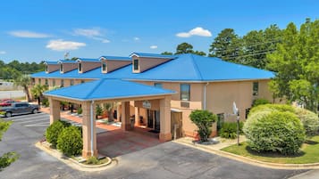 A hotel with a blue roof, covered parking, and a landscaped area with bushes and trees.