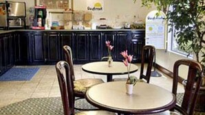 A kitchen area with dark cabinets, a coffee machine, and a sign that reads 'Quaker.' There are two round tables with chairs, and a potted plant on one table. - Gray Inn and Suites (Gray)