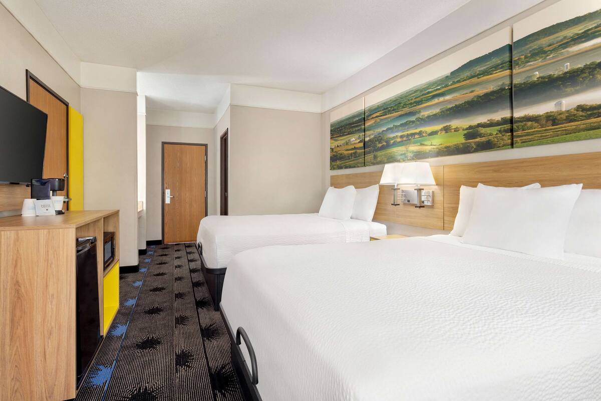 Ein Hotelzimmer mit zwei Betten, einem Schreibtisch, einem Flachbildfernseher, einer Kaffeemaschine und einem Landschaftsbild an der Wand.