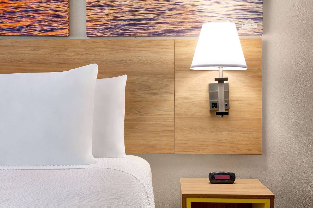 Ein Hotelzimmer mit einem Holzkopfteil, einem weißen Bett mit Kissen, einer Wandleuchte und einem kleinen Tisch mit einer Uhr.