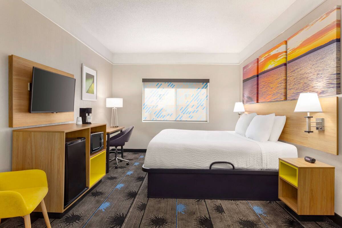 Ein Hotelzimmer mit Bett, Schreibtisch, Stuhl, Fernseher und einem Kunstwerk an der Wand.
