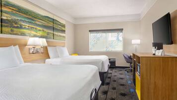 A hotel room with two beds, a desk, a TV, and a large window with blinds.