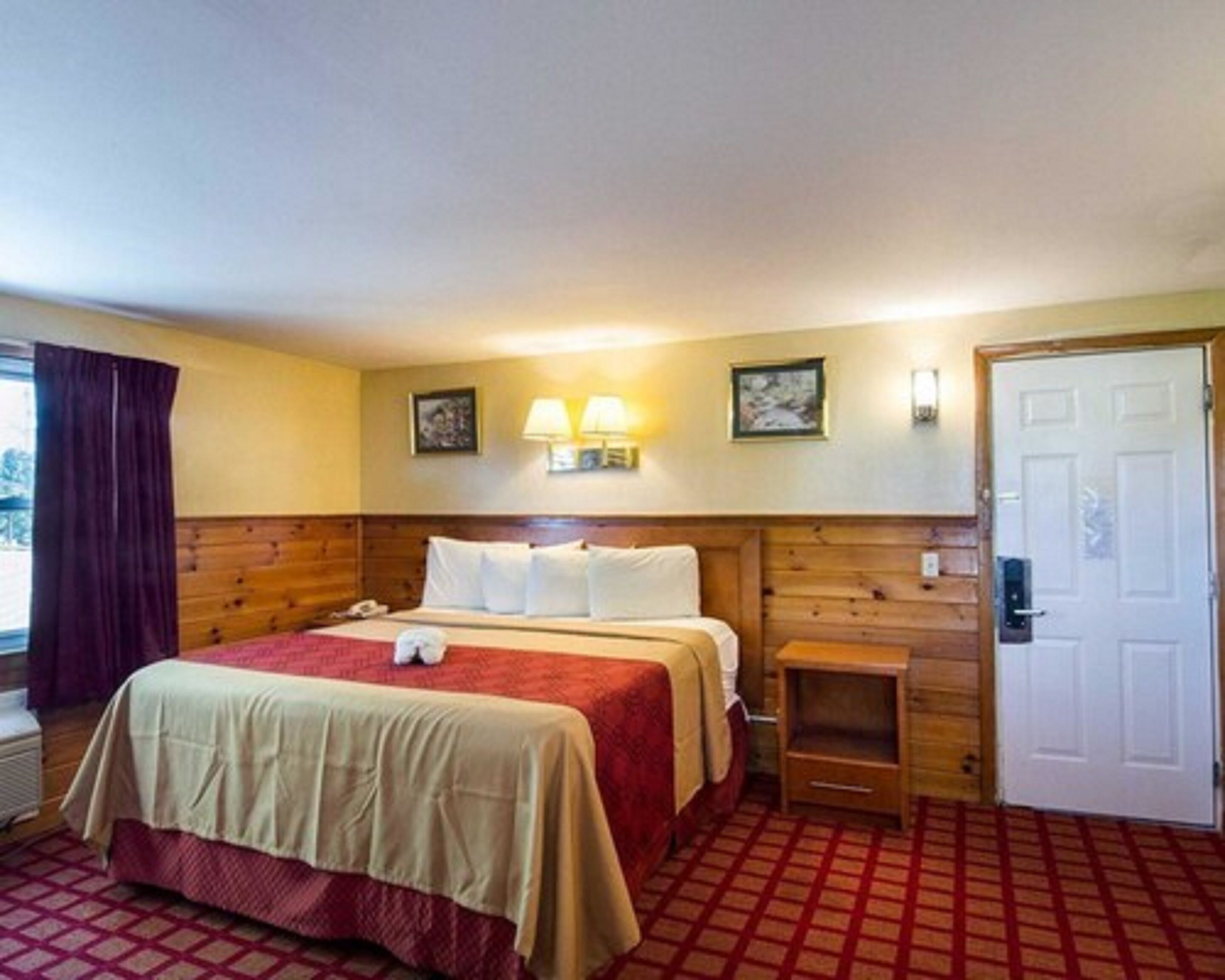 A hotel room with a wooden headboard, a bed with a red and beige bedspread, two wall-mounted lamps, a small wooden nightstand, and a door with a handle.