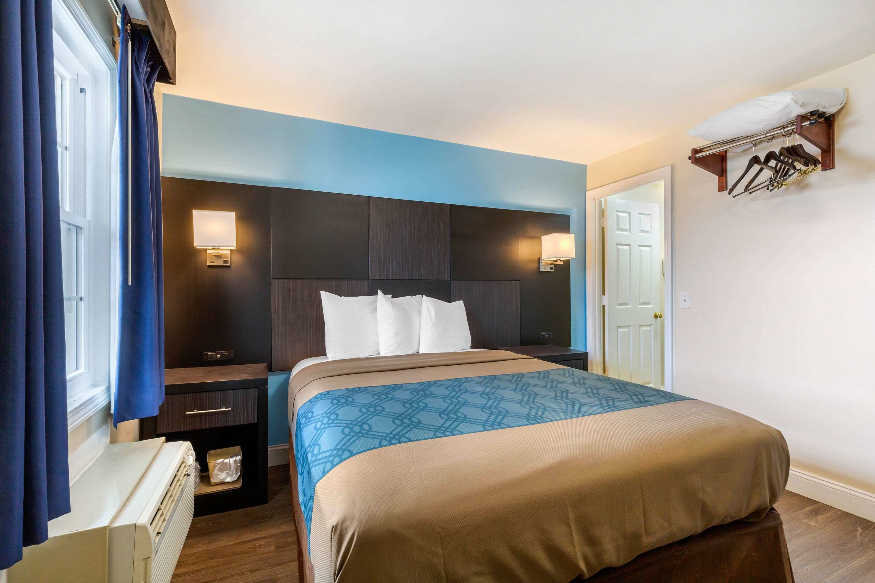 A hotel room with a bed, bedside tables, a nightstand, a lamp, a heater, a coat rack, and a door.