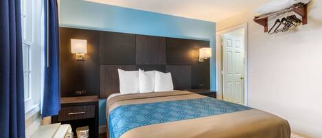 A hotel room with a bed, bedside tables, a nightstand, a lamp, a heater, a coat rack, and a door.