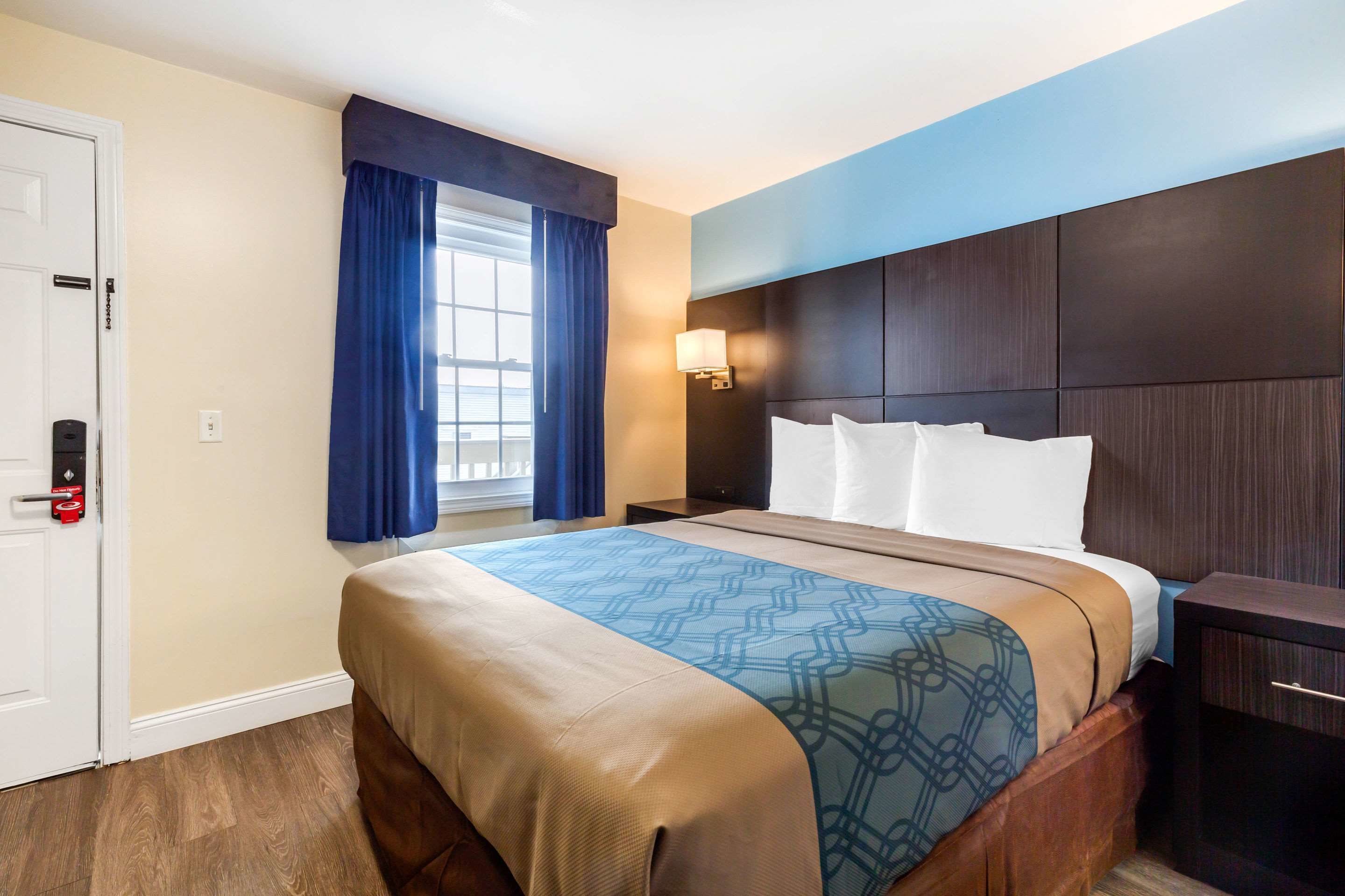 Habitación de hotel con una cama grande, una mesita de noche, una ventana con cortinas y una puerta con cerradura.
