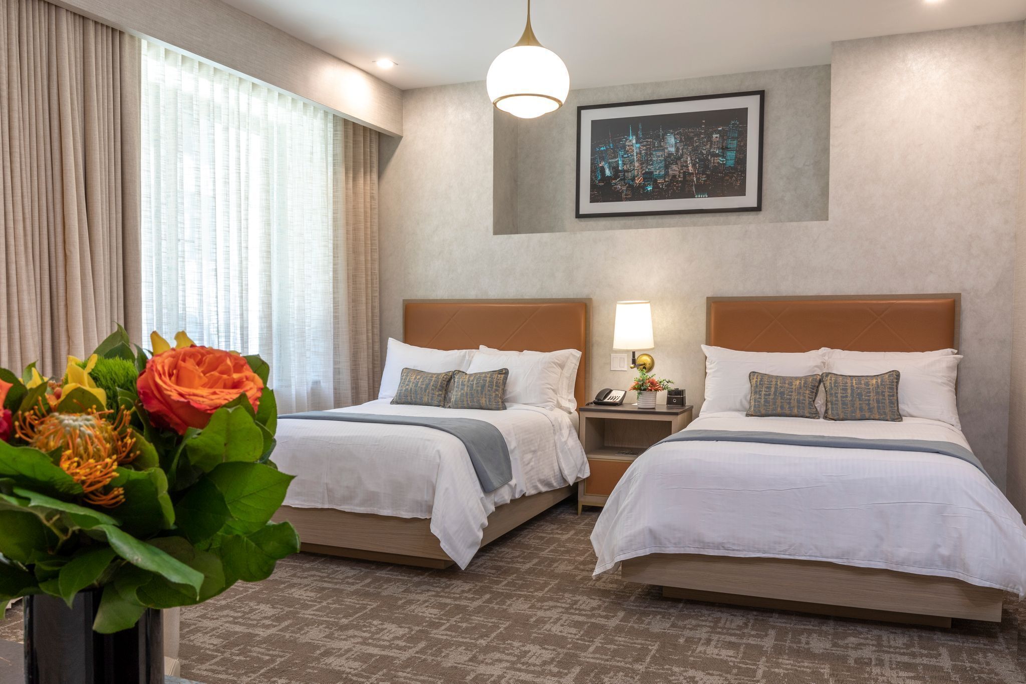A hotel room with two beds, a nightstand with a lamp, a framed cityscape picture on the wall, and a vase of flowers on the left.