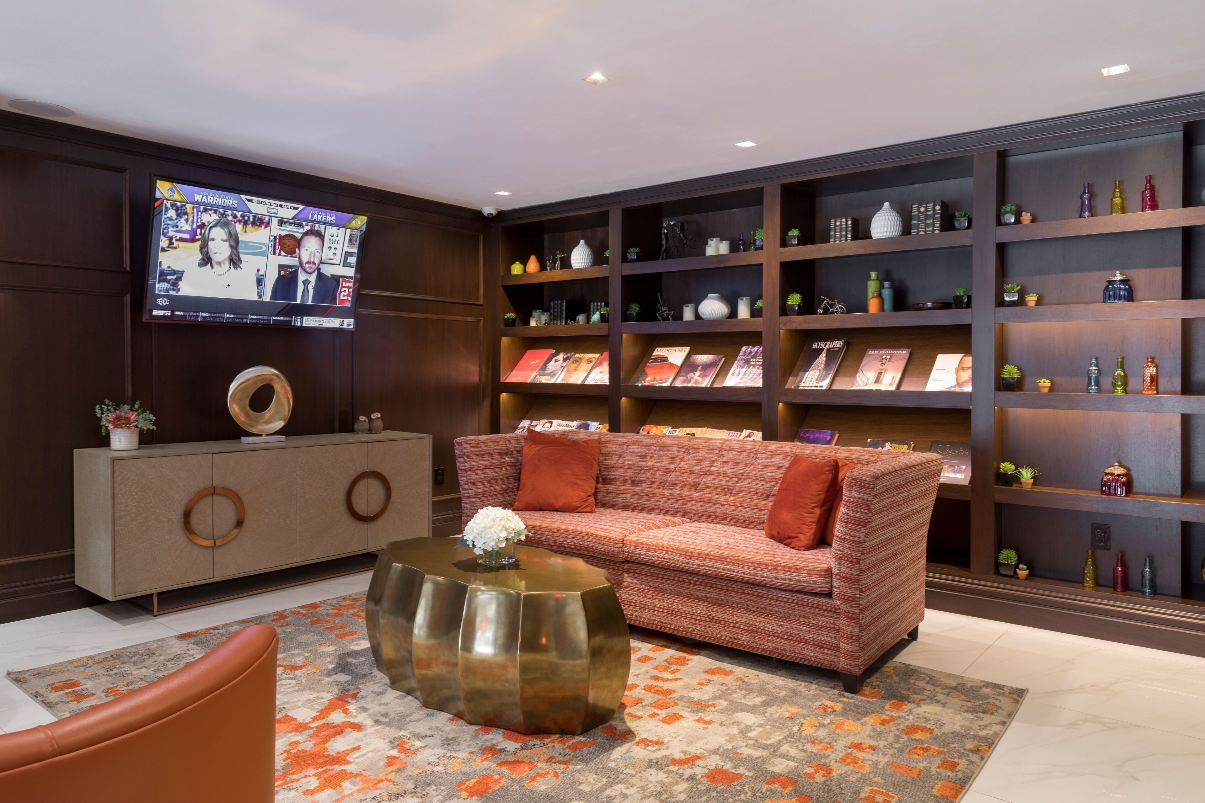 A modern living room with a sofa, coffee table, and a TV displaying a sports game. There are shelves with decorative items and books.