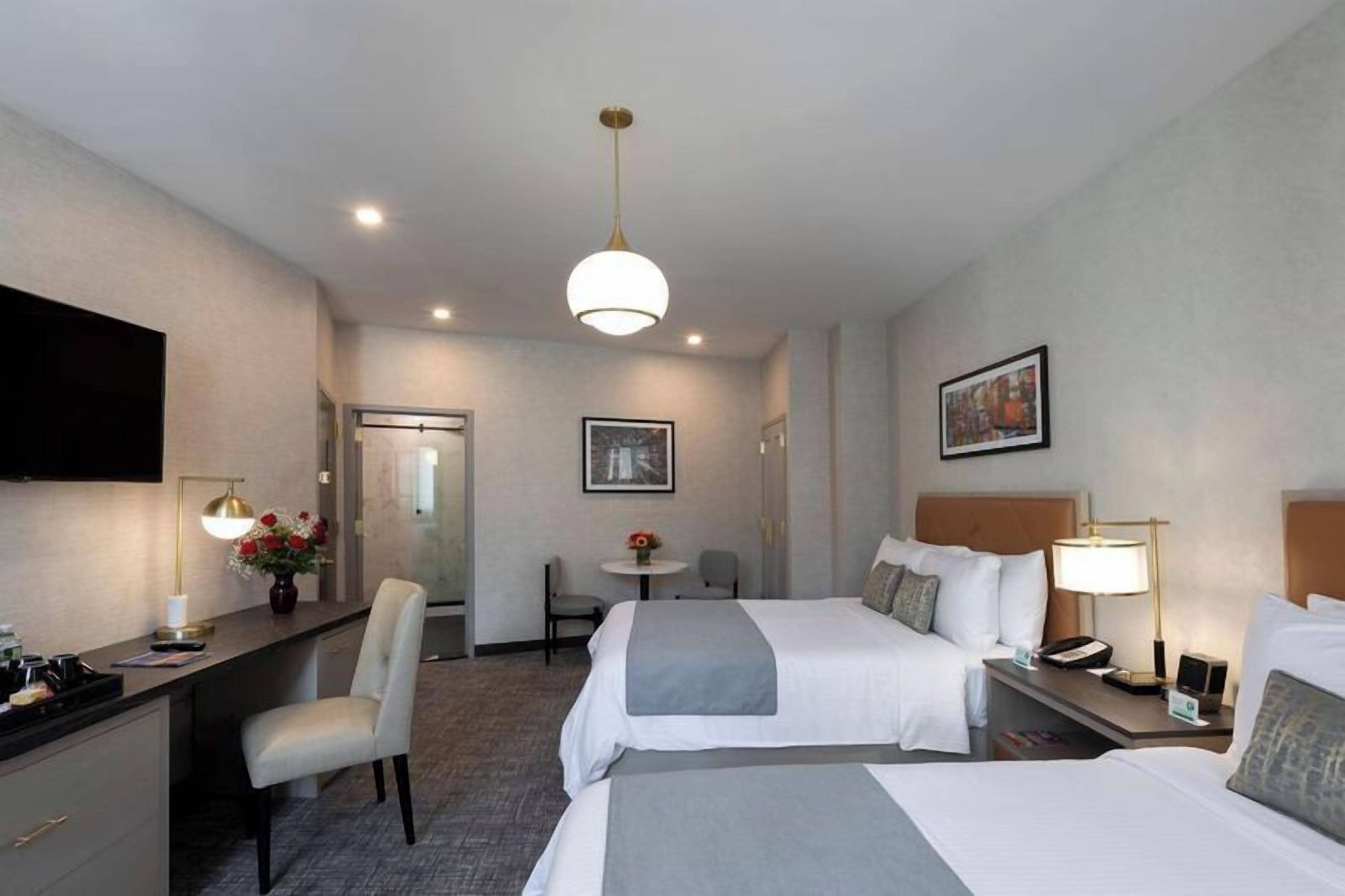 Une chambre d’hôtel avec deux lits, un bureau, une chaise, une télévision et une petite table sur laquelle est posé un vase de fleurs.