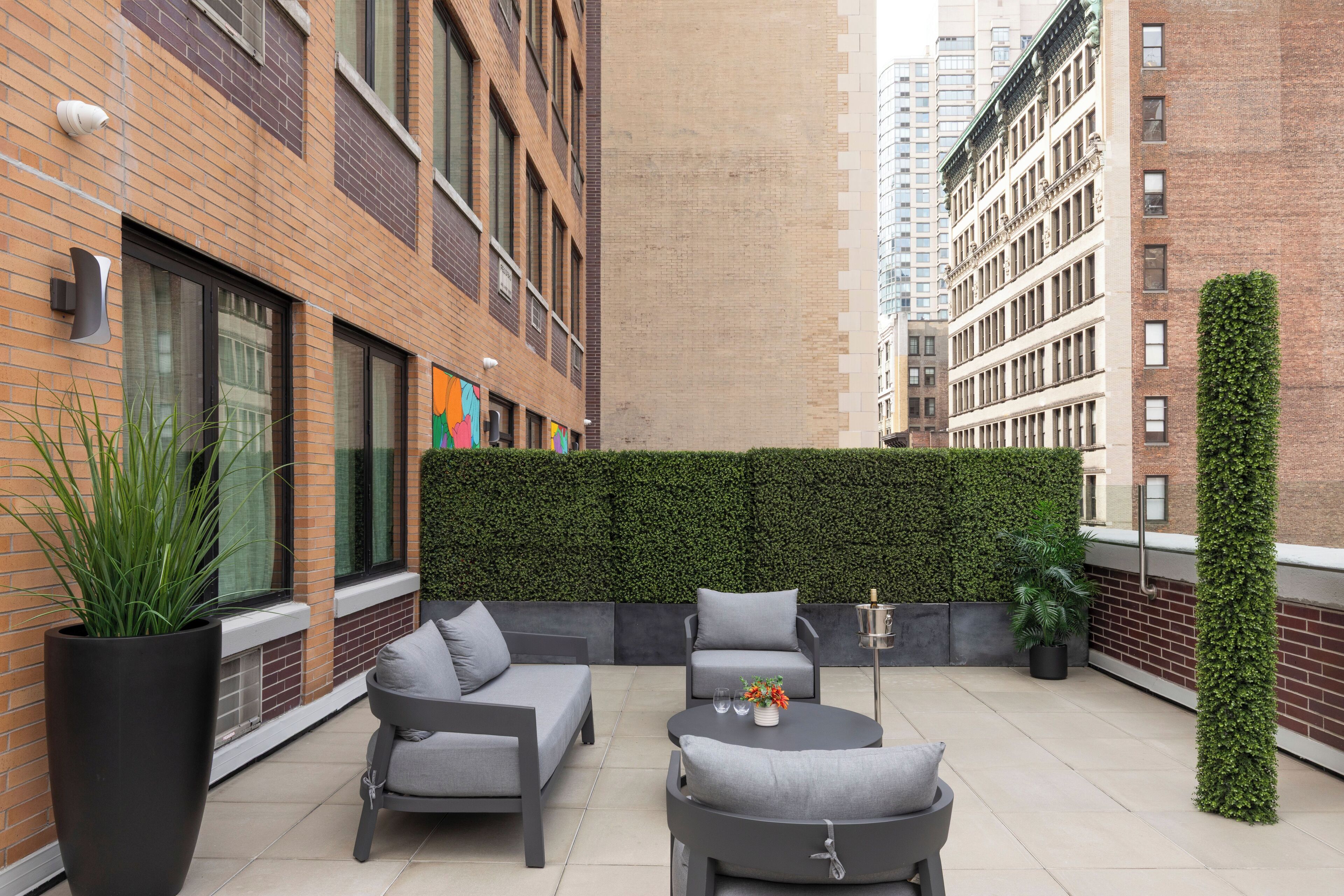 A rooftop patio with outdoor seating, a green wall, and tall buildings in the background.