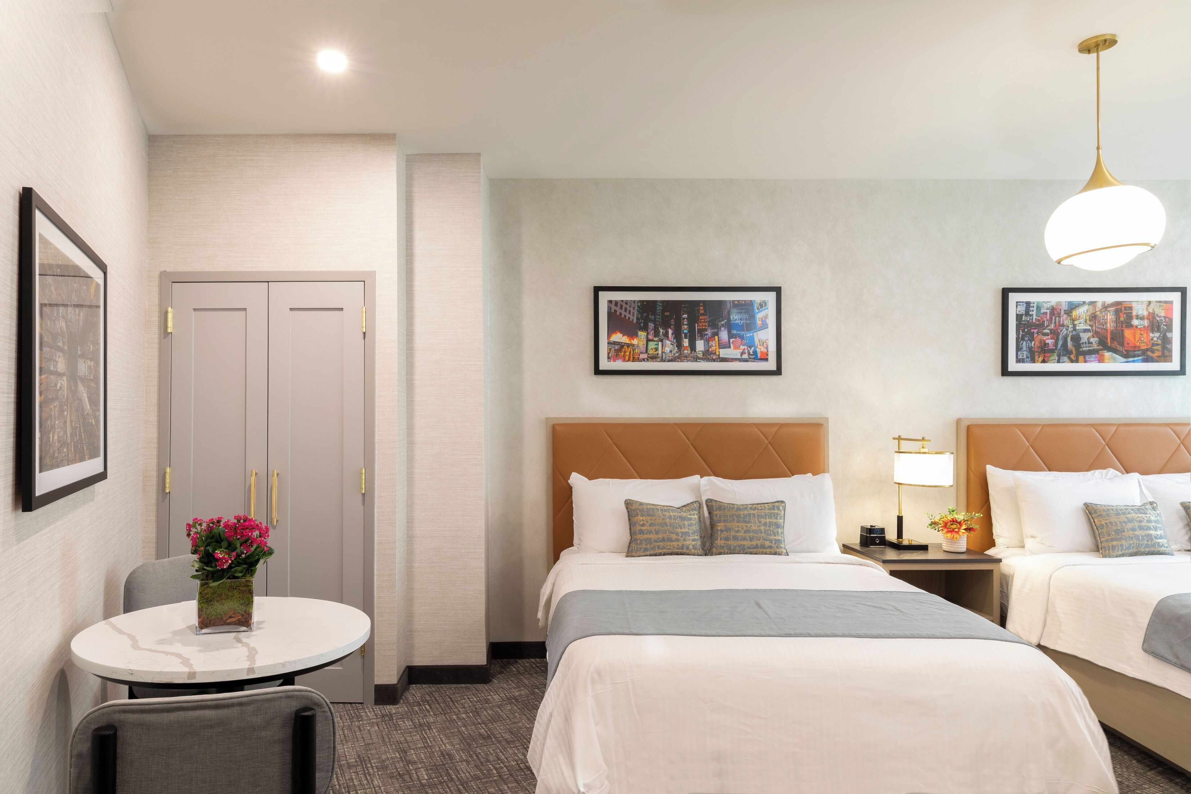 Ein Hotelzimmer mit zwei Betten, einem runden Tisch mit Blumenvase, einem gerahmten Bild an der Wand und einer Lampe auf dem Nachttisch.
