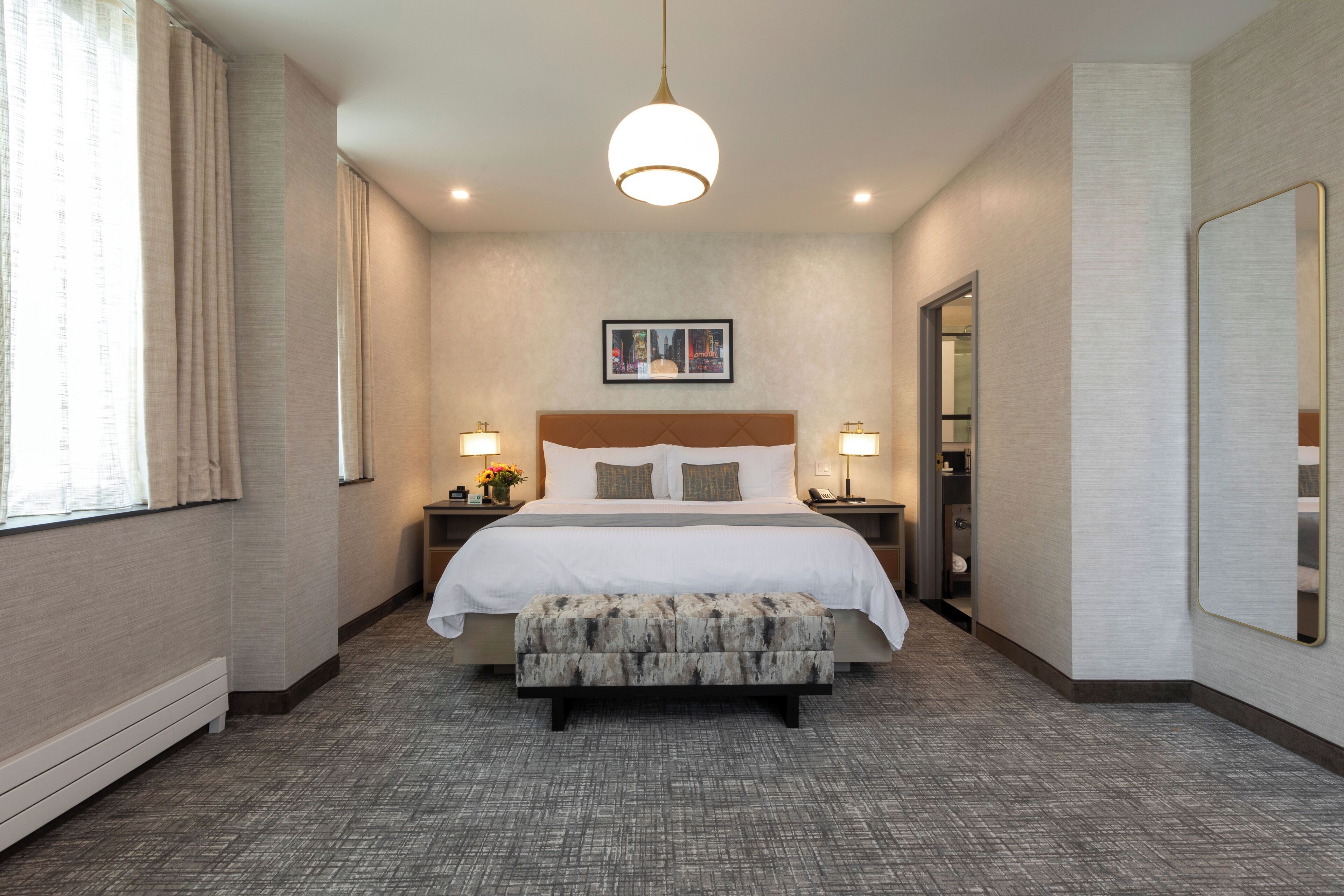 A hotel room with a large bed, a bedside table with a lamp, a bench, a framed picture on the wall, and a mirror on the wall.