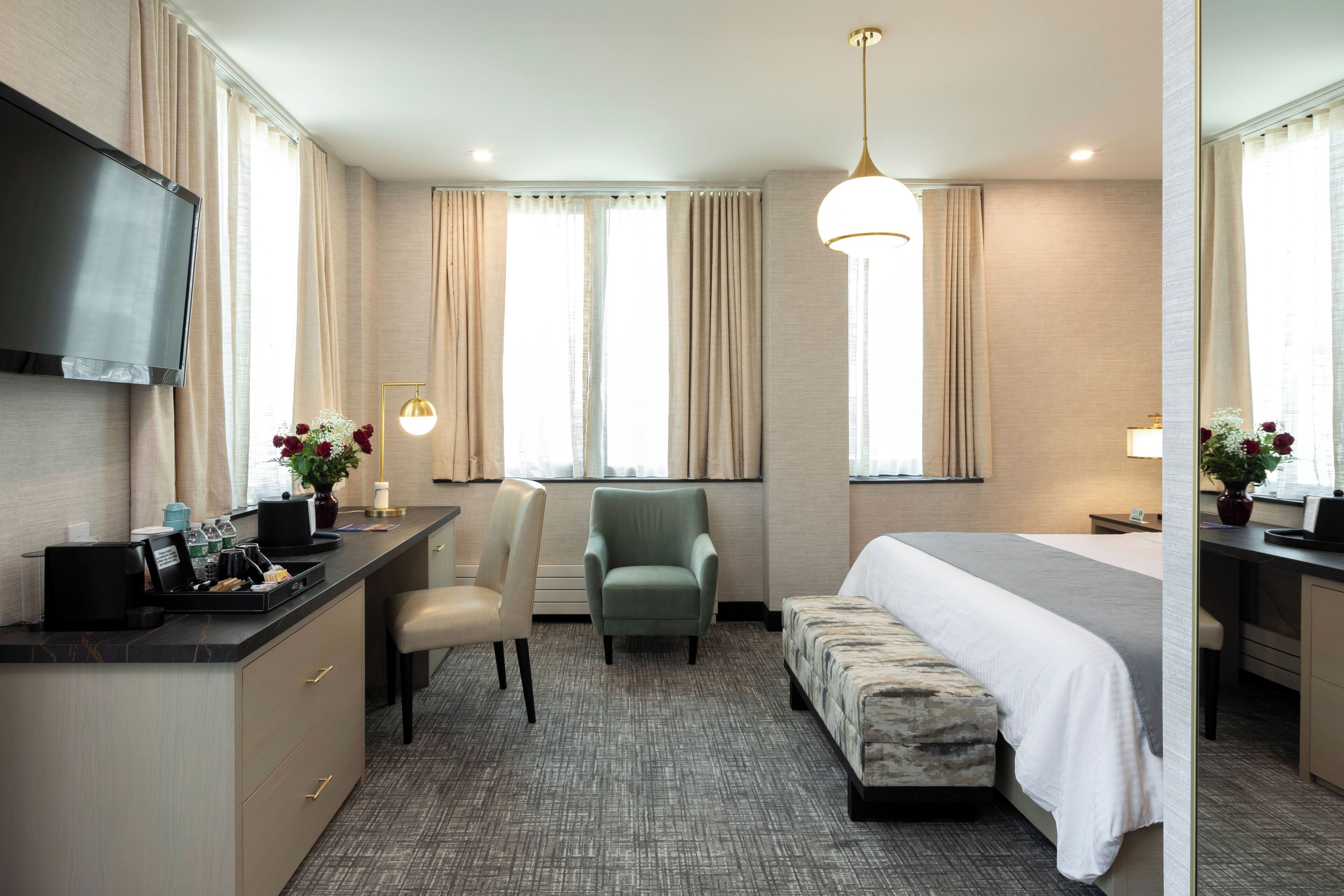 A hotel room with a bed, a desk with a flat-screen TV, a chair, a sofa, and a large window with curtains.