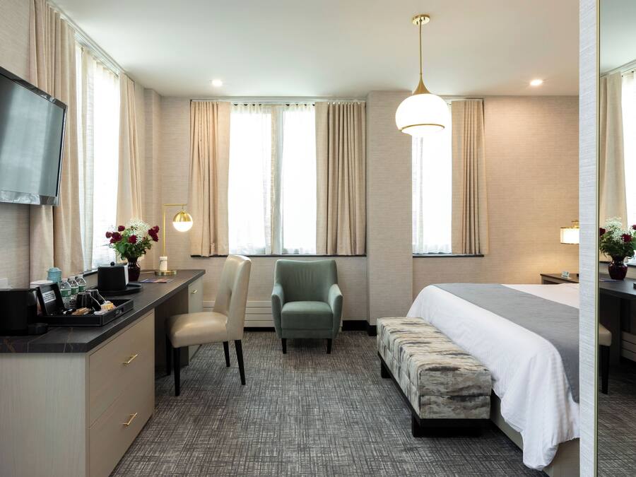 A hotel room with a bed, a desk with a flat-screen TV, a chair, a sofa, and a large window with curtains.
