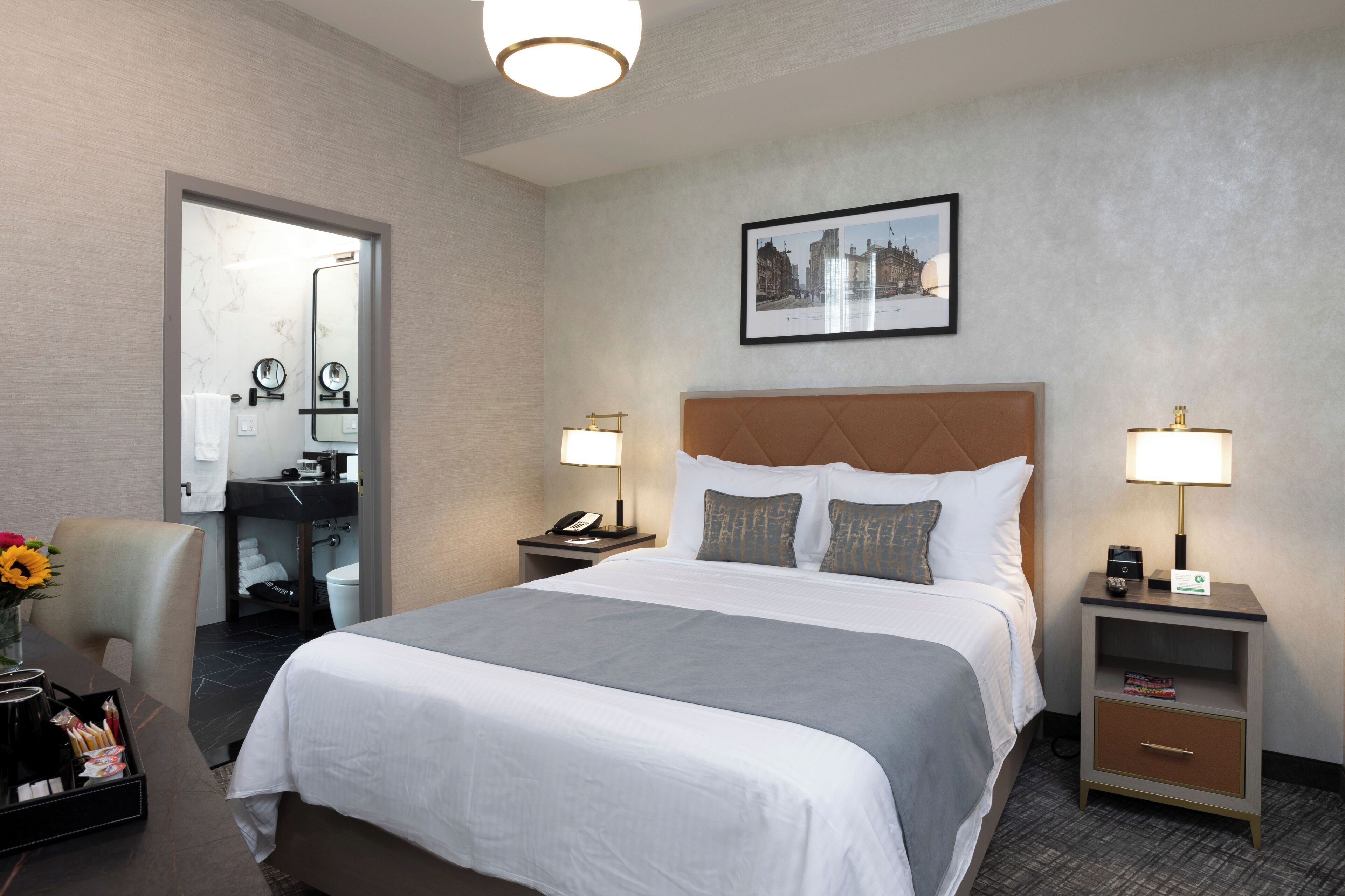 A hotel room with a large bed, two bedside tables with lamps, a desk with a chair, and a bathroom visible through an open door.