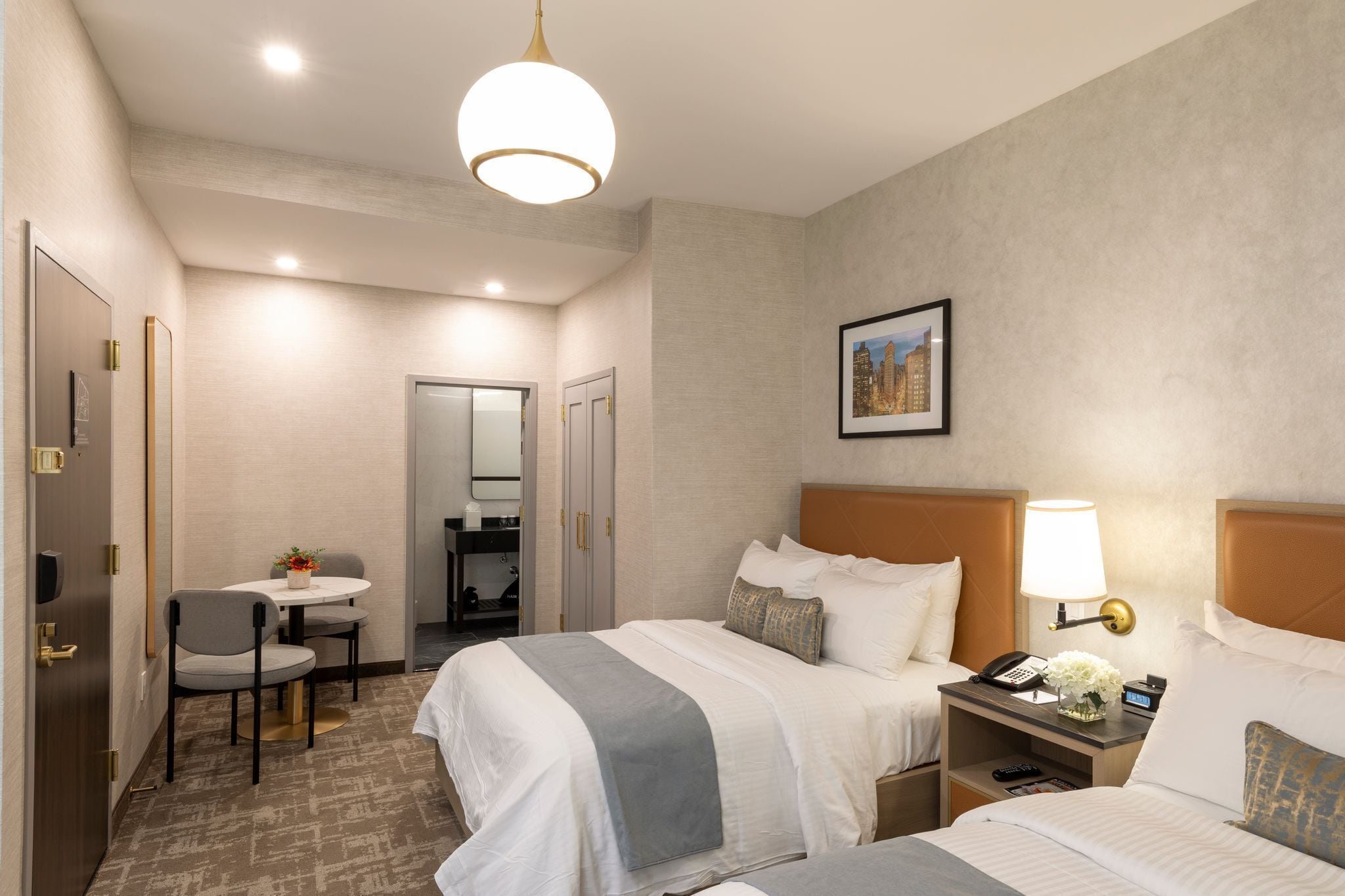 A hotel room with two beds, a desk with a chair, a small table with flowers, a lamp, and a framed picture on the wall.