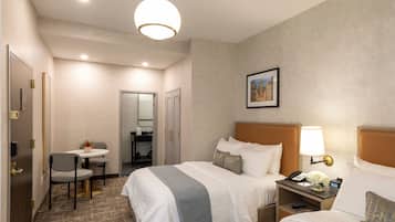A hotel room with two beds, a desk with a chair, a small table with flowers, a lamp, and a framed picture on the wall.